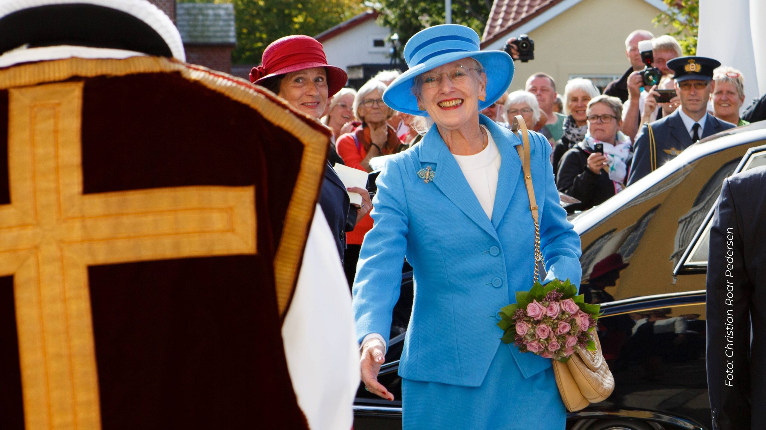 Dronning Margrethe klædt i blåt med en lyserød buket blomster i hånden hilsende på en præst, der ses fra ryggen. I baggrunden ser en folkemængde på.