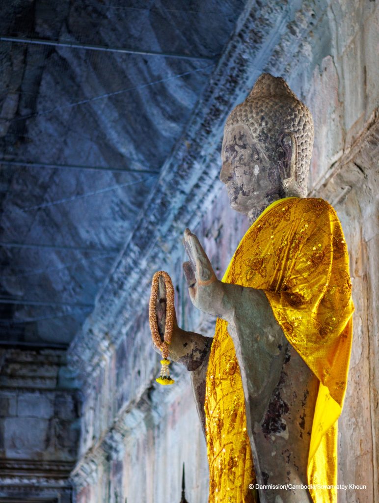 En gudestatue i et tempel. Klædt i en gylden dragt og holder en kæde i sin hånd.
